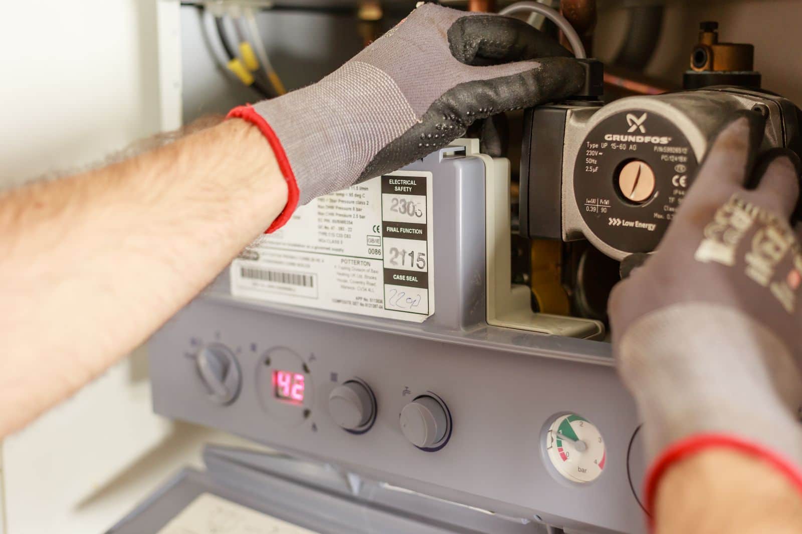 Plumber's gloved hands servicing a wall-hung water heater, temperature display reading 142