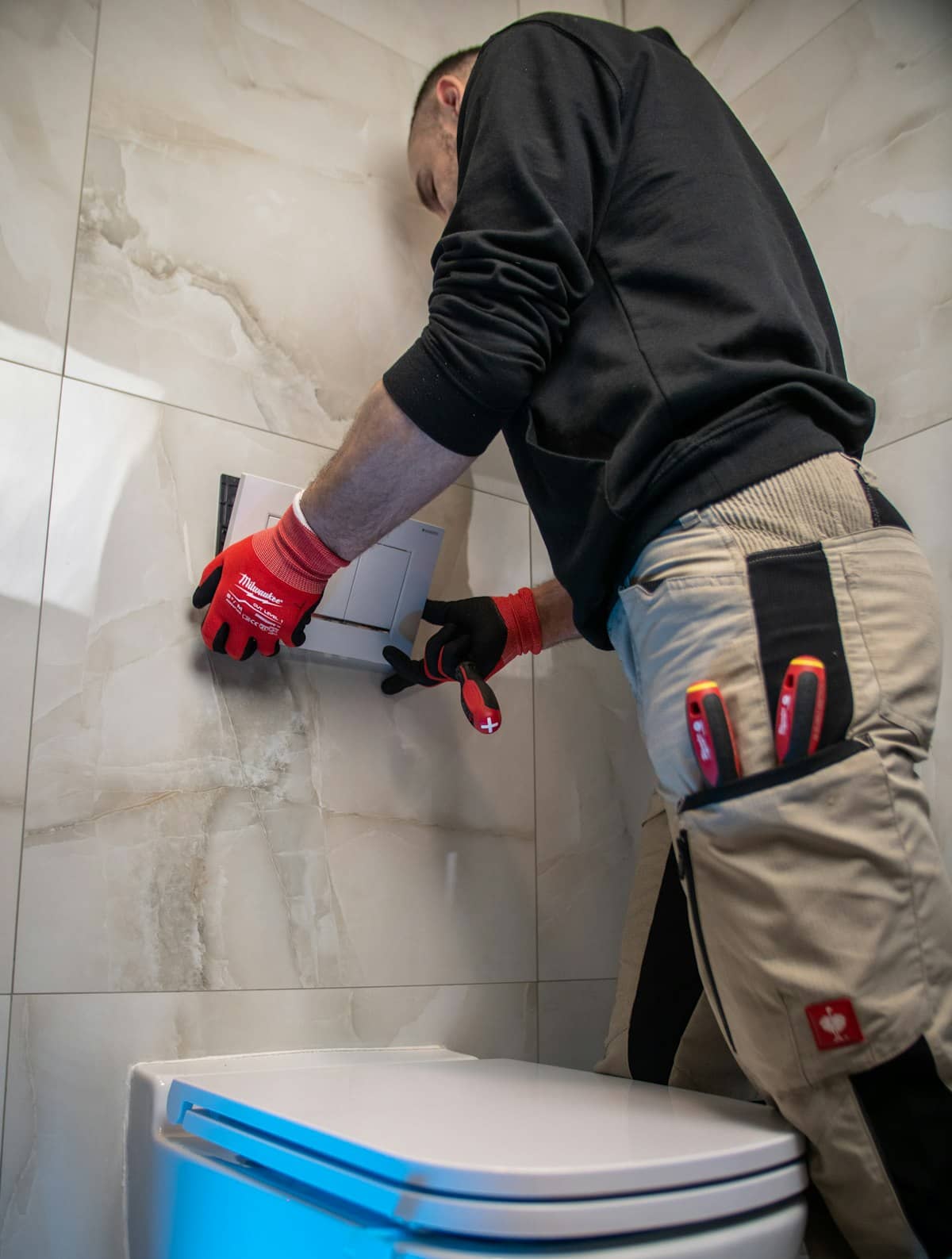 Plumber in work gloves installing a wall-mounted flush plate in a tiled bathroom