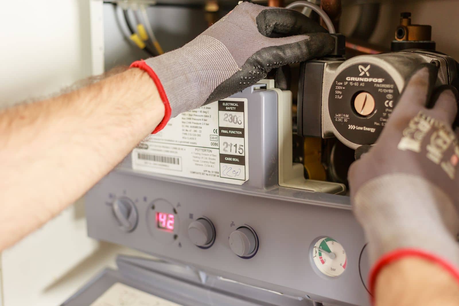 Plumber's gloved hands servicing a wall-hung water heater, temperature display reading 142